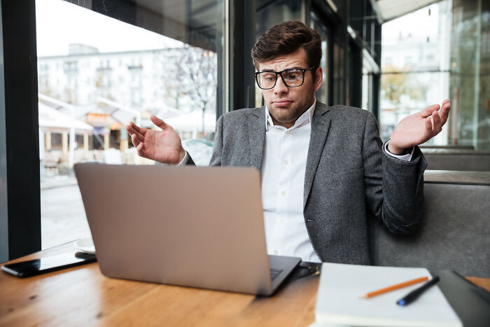 Young man with glasses shrugging in front of laptop, portraying mind-boggling conversations with adults and confusion.