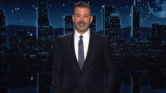 Jimmy Kimmel standing on a nighttime city skyline set, wearing a dark suit and striped tie during a TV broadcast.