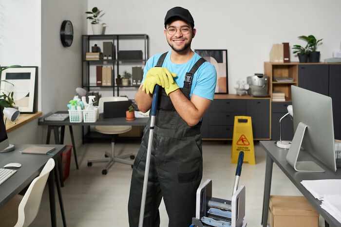 Young male janitor wearing gloves and overalls, smiling while holding mop in a modern office, highlighting school incidents cleanup.
