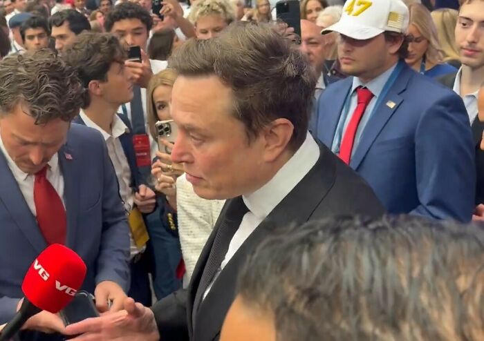 Elon Musk in a crowd at an event, wearing a black suit and white shirt, amidst people recording on phones.