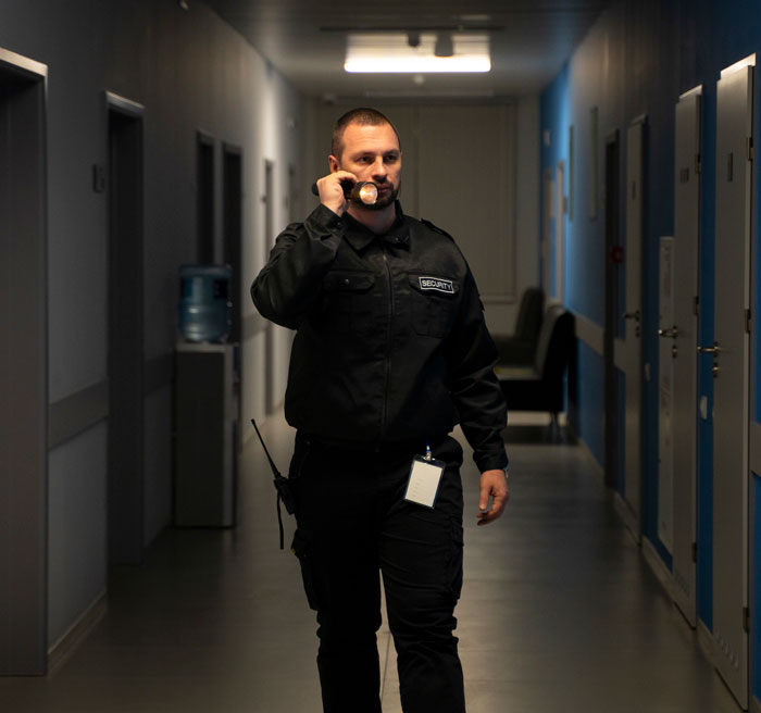 Security guard walking through a dimly lit hallway holding a flashlight, on a nighttime patrol during his job shift.