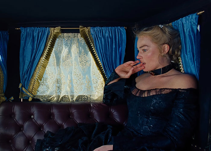 Margot Robbie in 1800s attire sitting in a vintage carriage with blue curtains, evoking Wuthering Heights atmosphere.