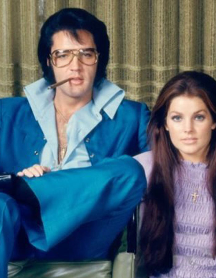 Elvis Presley in a blue suit smoking a cigar, sitting next to Priscilla Presley in a lavender top, vintage photo.