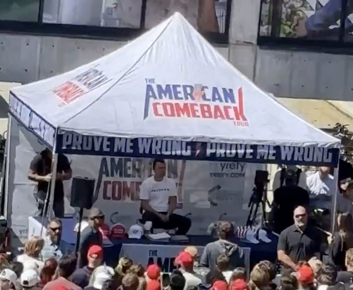Crowd gathered near commentator booth with The American Comeback tent and people wearing red hats at outdoor event.