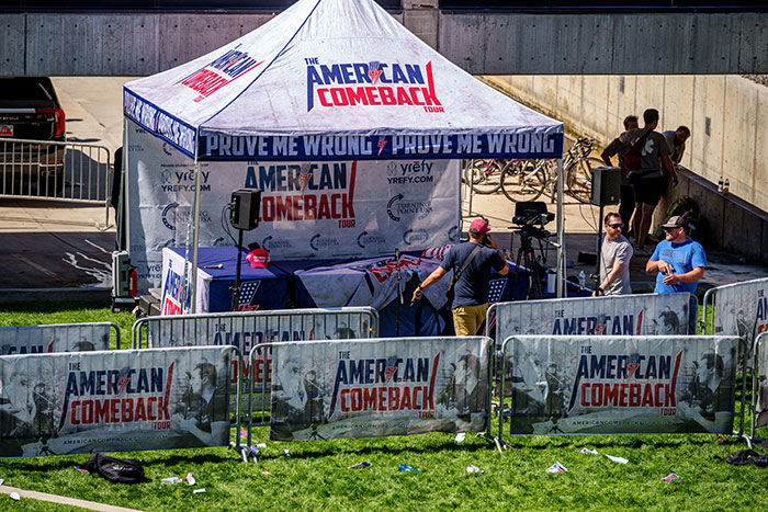 Outdoor tent setup with American Comeback Tour banners and people setting up recording equipment near metal barricades.