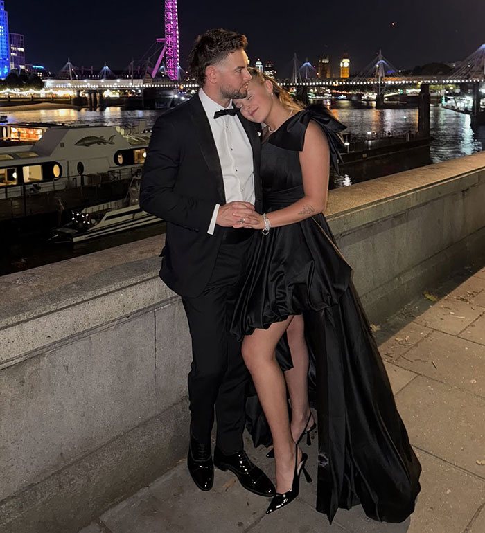 JoJo Siwa posing by the river at night, wearing a black dress, with a man in a tuxedo beside her.