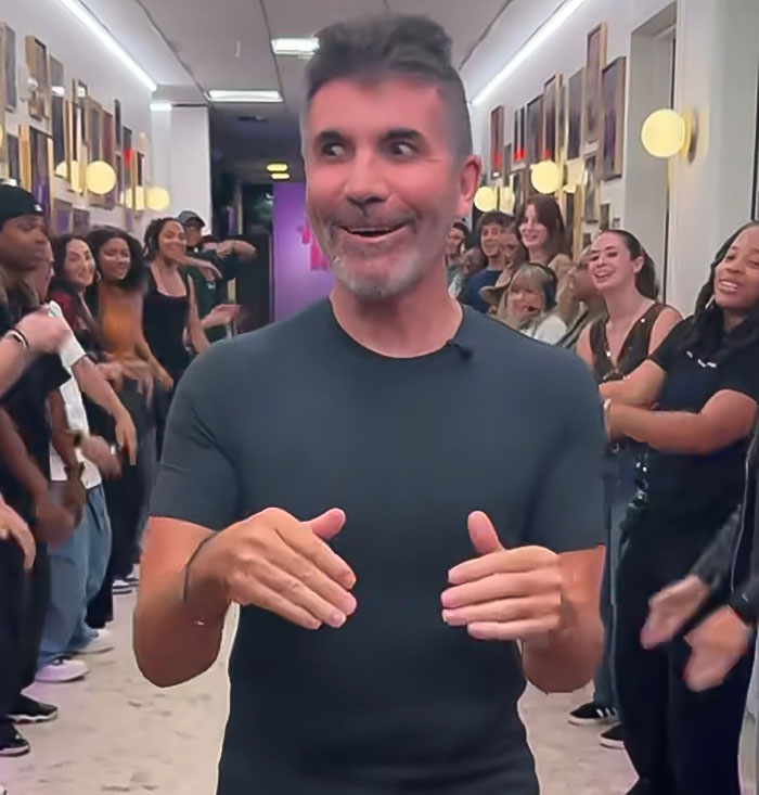 Simon Cowell smiling and gesturing while surrounded by a group of people in a hallway after bike accident recovery.