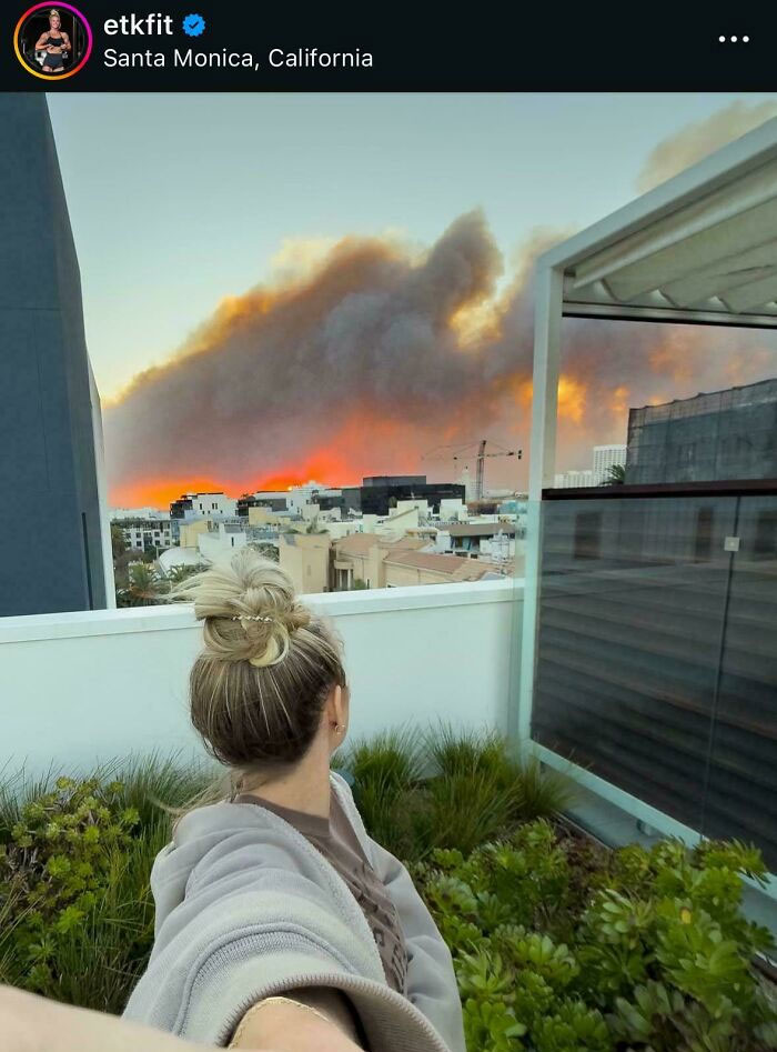 Fitness influencer taking a selfie outdoors with urban buildings and a large smoky sky in the background.