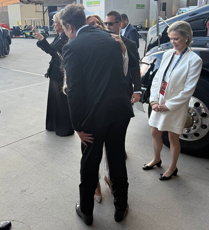 Elon Musk in a black suit interacting with people outdoors near vehicles amid event attendees in formal attire.