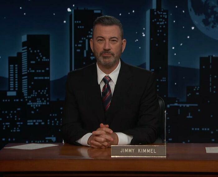 Jimmy Kimmel in a suit seated at a desk with a cityscape background, related to staff mutiny and tearful return. Jimmy Kimmel in a suit seated at a desk with a cityscape background, related to staff mutiny and tearful return.