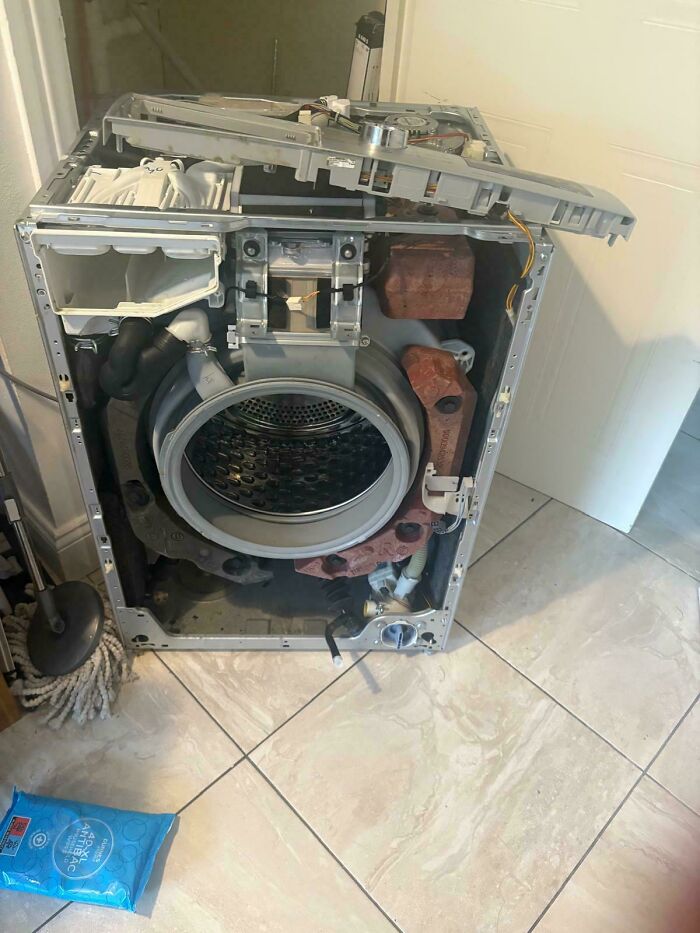 Partially disassembled washing machine placed on tiled floor, illustrating the impact of consumerism on product durability.