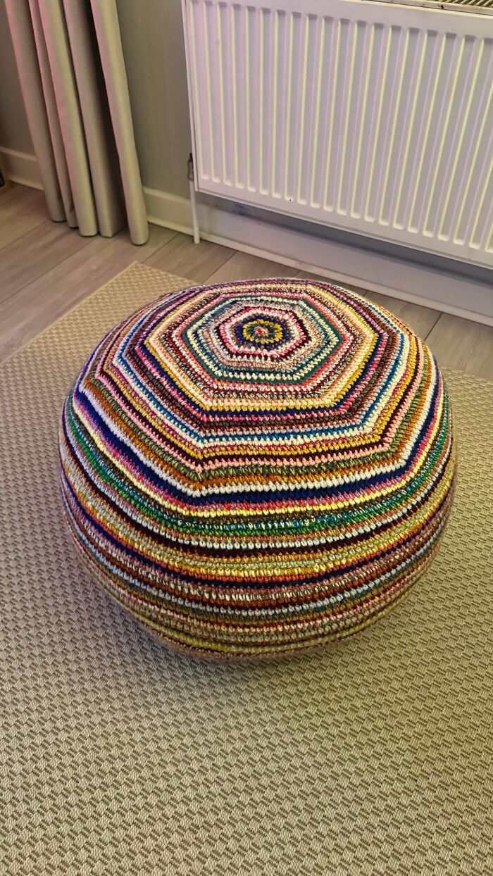 Colorful crocheted pouf with vibrant striped pattern placed on a textured beige rug in a cozy room setting