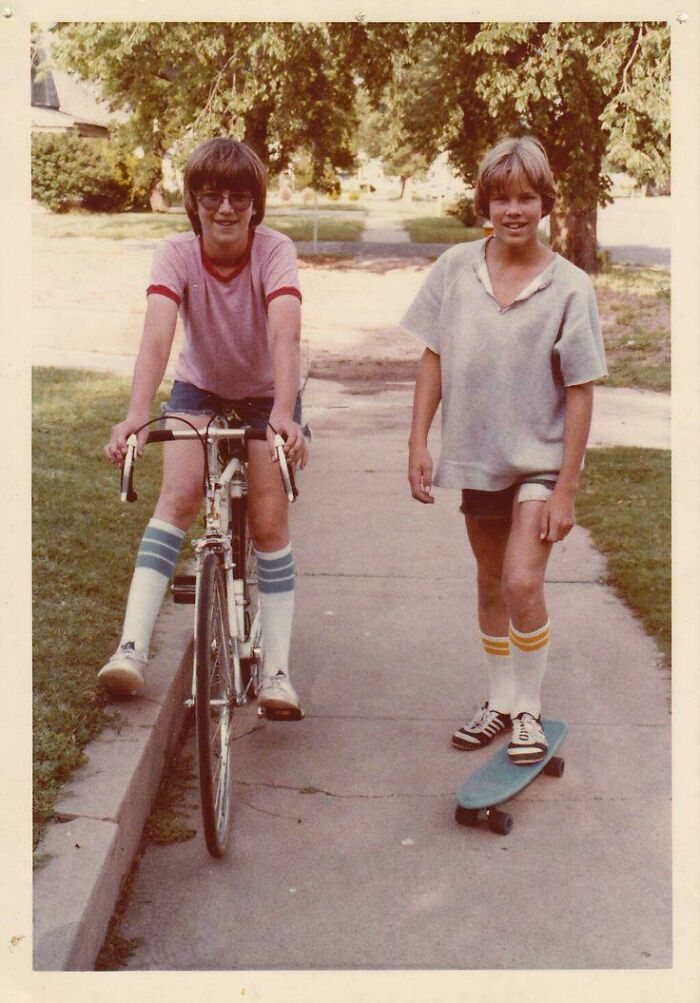 Two kids from the past riding a bike and skateboard on a sidewalk, nostalgic photo to make you feel old as heck.