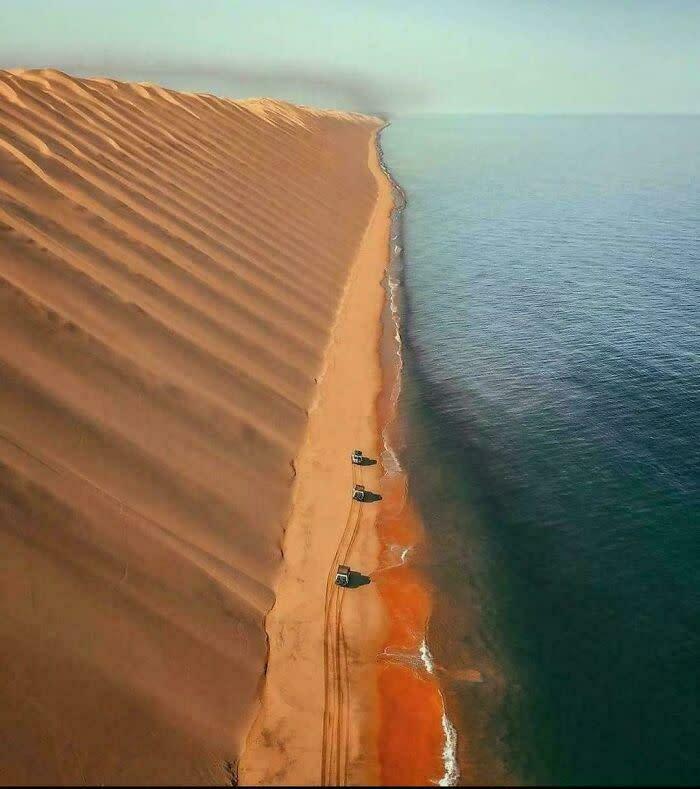 An aerial view of extremely large sand dunes meeting the ocean, with vehicles on the beach, a beautiful but eerie landscape.