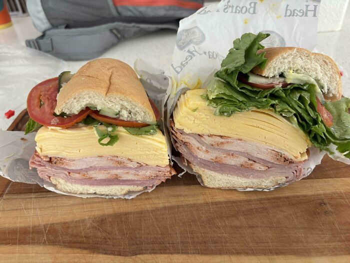 Two unusual sandwiches with thick slices of cheese and deli meat stacked high on a wooden board, showing odd dish layering.