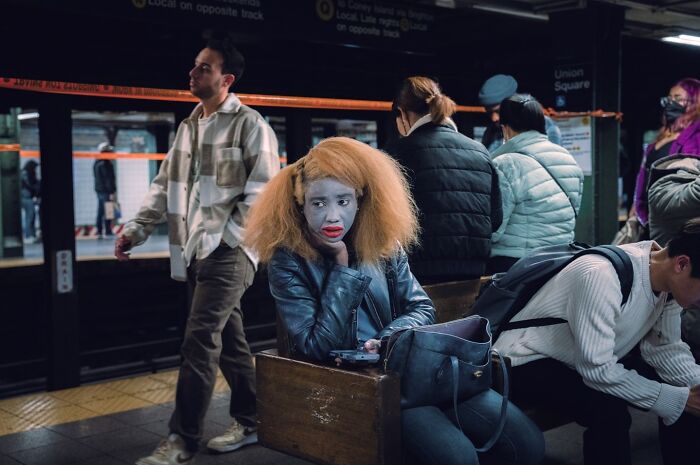 Woman with striking makeup and large hair sitting on a subway bench among commuters in a cinematic street photo.