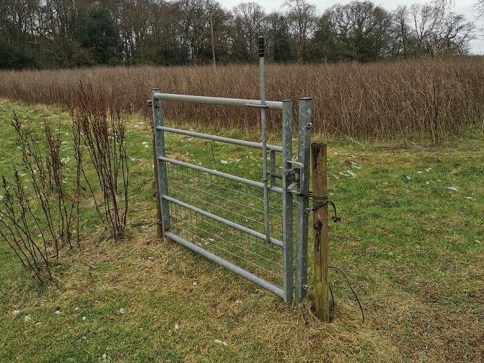 Half-installed metal farm gate awkwardly attached to a lone wooden post in a grassy field, showing hilariously bad job.