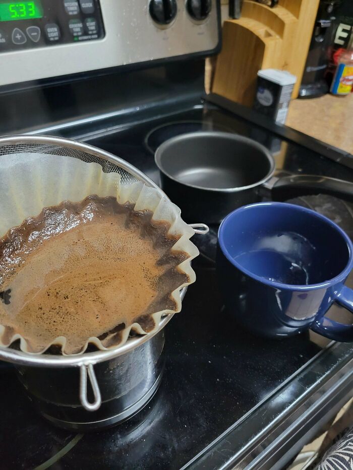 Kitchen stovetop with coffee brewing in a homemade filter setup next to a blue mug, illustrating consumerism and simplicity.