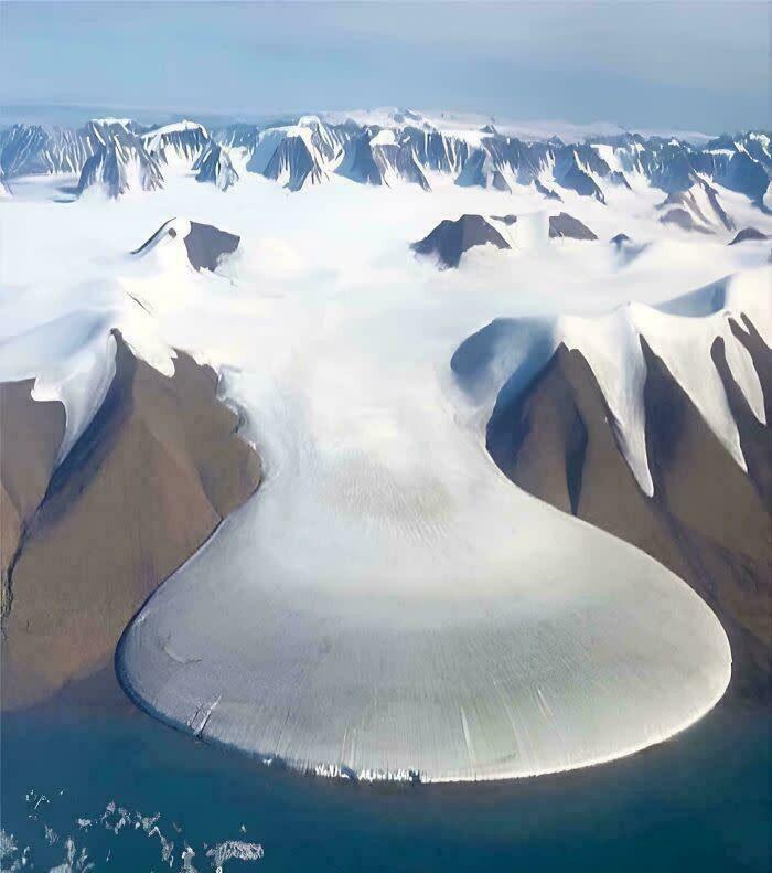 An aerial view of an extremely large, eerie glacier meeting the ocean, surrounded by snow-capped mountains.