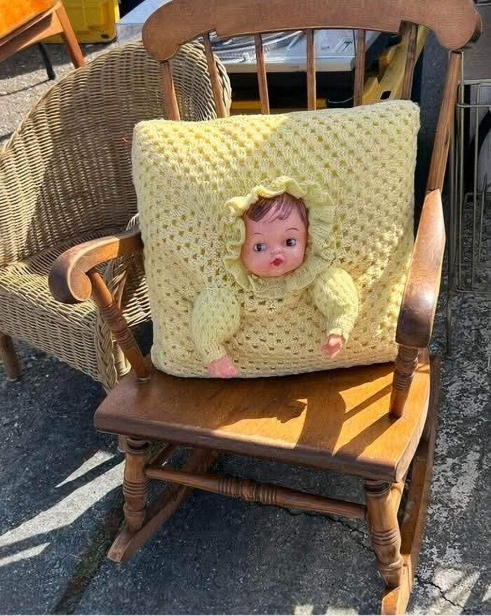 Crocheted yellow pillow with a doll face and arms, an example of horrendous DIY craft shaming projects on a wooden chair.