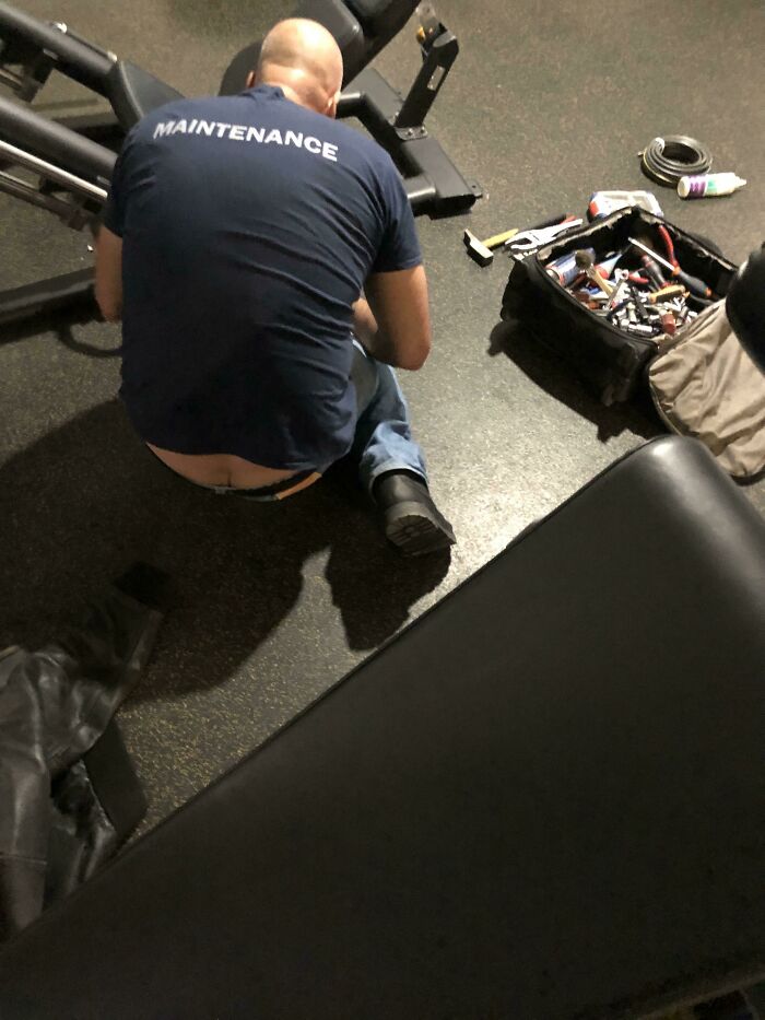 Man wearing a maintenance uniform exposing lower back while working with tools on a gym machine, funny uniform moment.