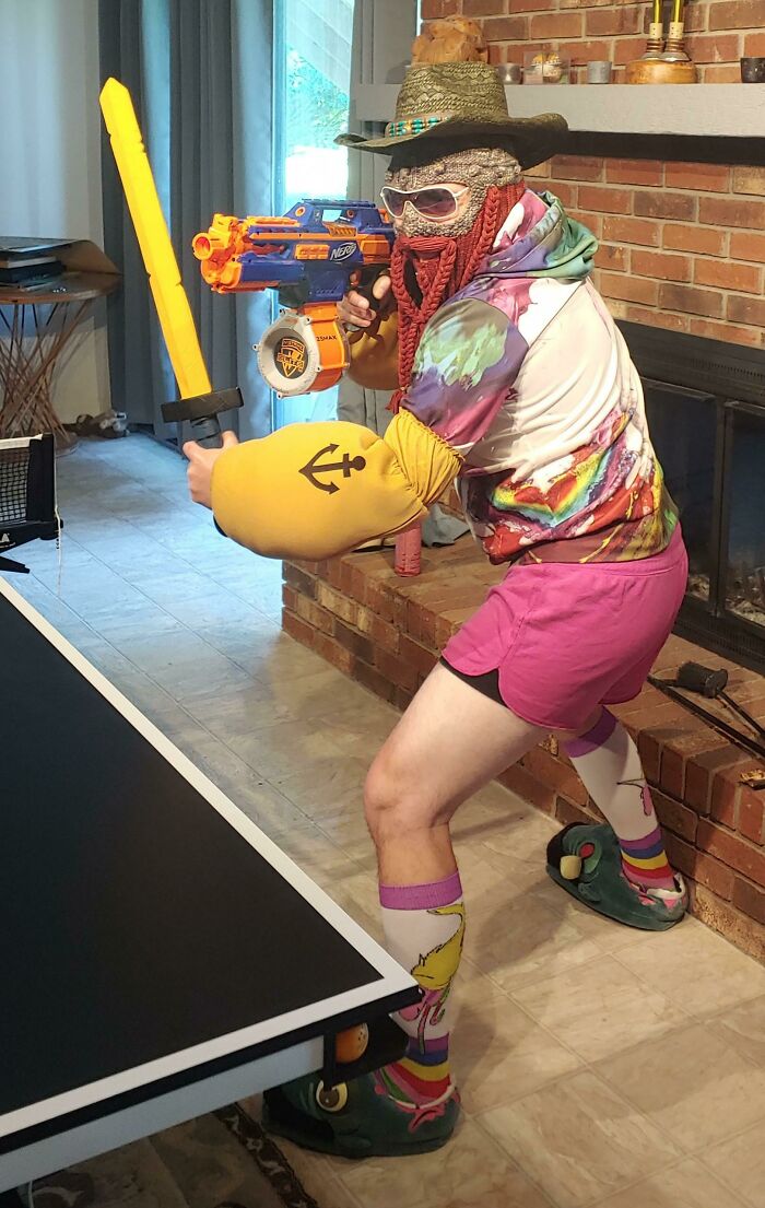 Person wearing a funny uniform with a fake beard, oversized arm, toy sword, and Nerf gun in a playful indoor setting.