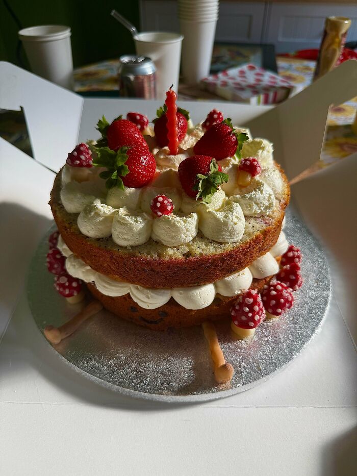 Two-layer baked cake decorated with whipped cream, strawberries, and small red mushroom-shaped toppings on a silver board.