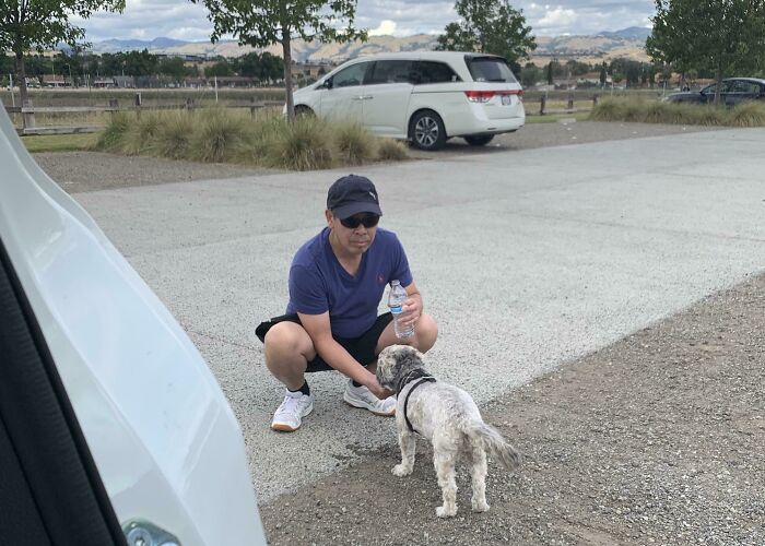 Dad wearing sunglasses and cap crouching to pet a small dog in a parking lot, showing softened attitude toward pets.