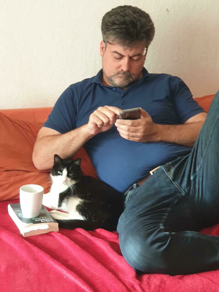 Man in a blue shirt relaxing on a couch with a black and white cat, showing dads becoming total softies with pets.