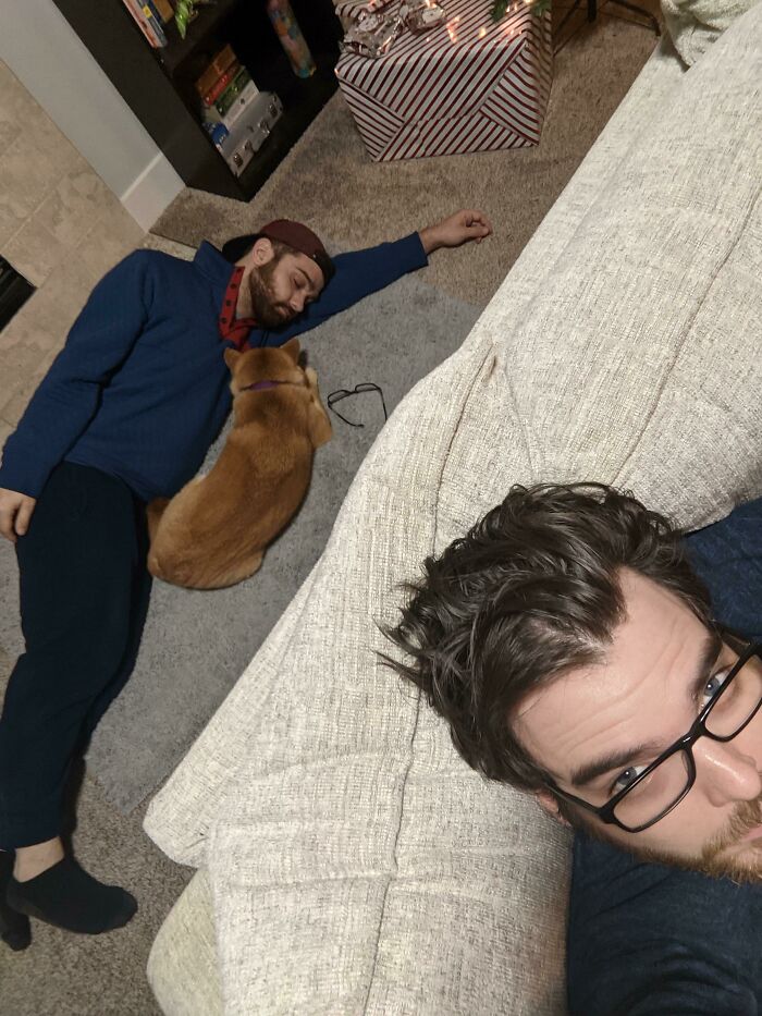 Man lying on the floor next to a dog, while another man with glasses relaxes on a beige couch in a cozy living room.