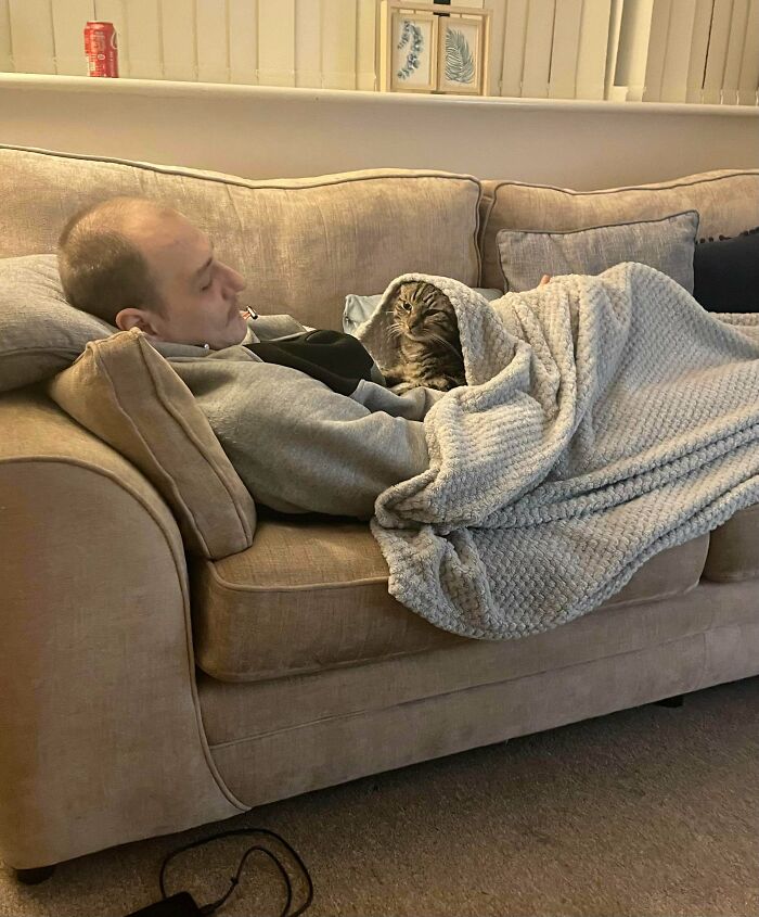 Man on couch covered with blanket, cuddling a cat, showing dads who said no pets becoming total softies.