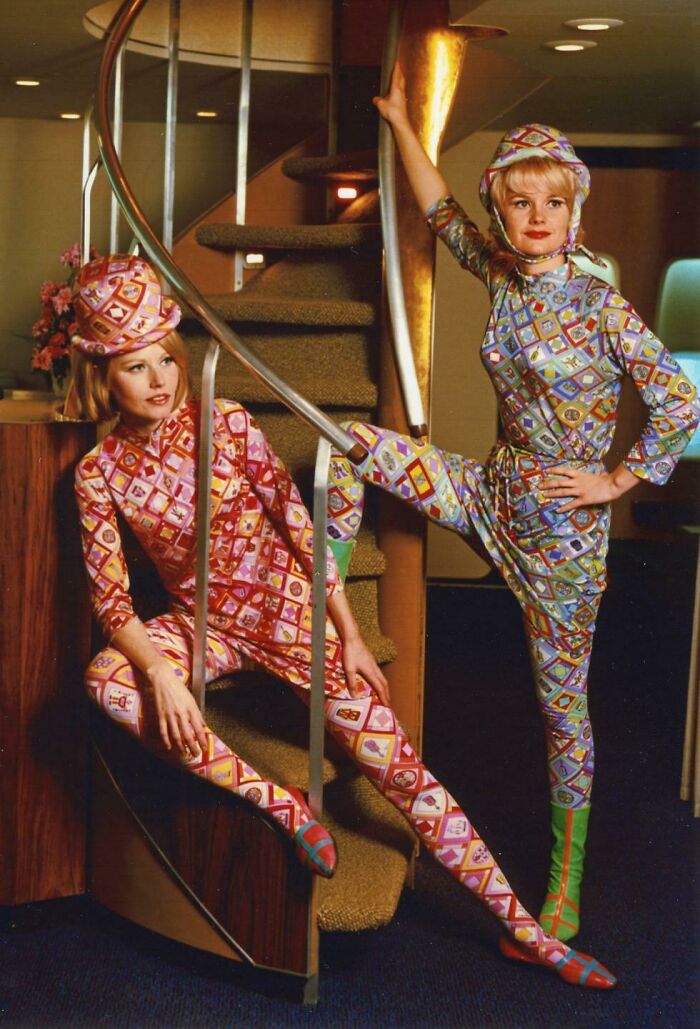 Two women wearing colorful retro patterned uniforms posing on and near a spiral staircase in a vintage setting.