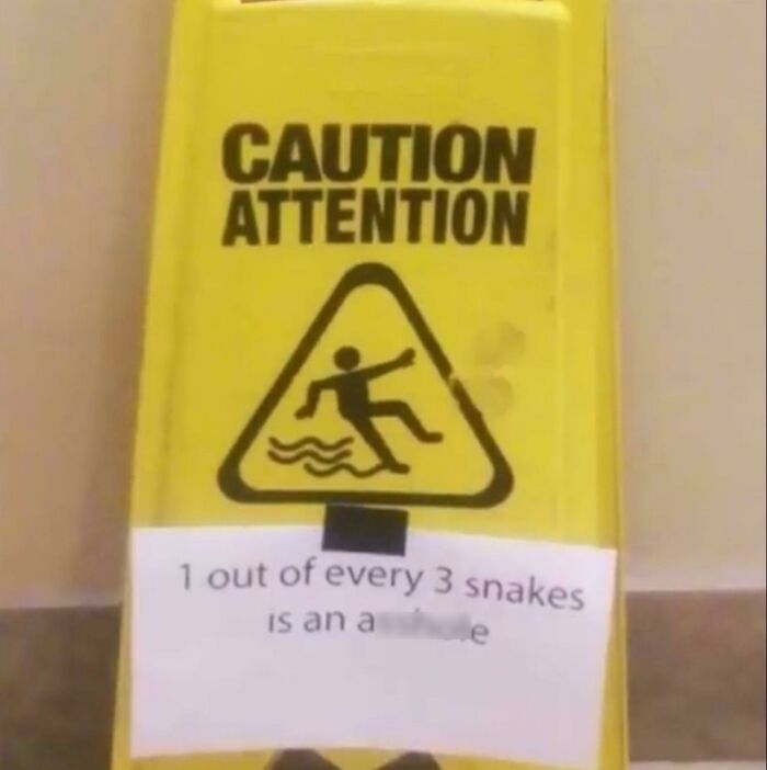 Yellow caution sign vandalized with a humorous note about snakes, showcasing creative public property vandalism.