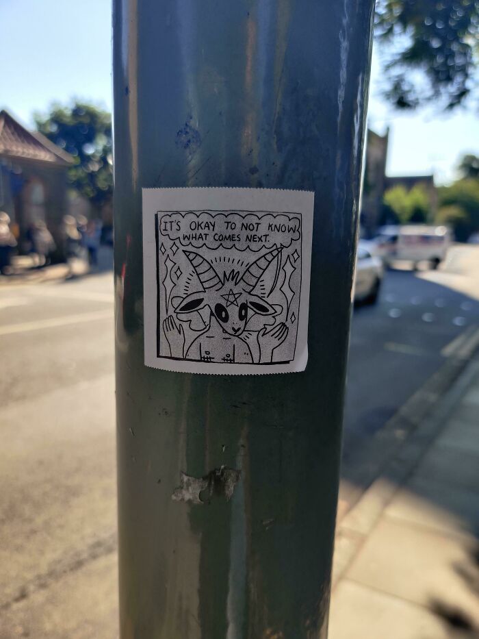Sticker vandalism on a street pole shows a cartoon goat with the message it's okay to not know what comes next.