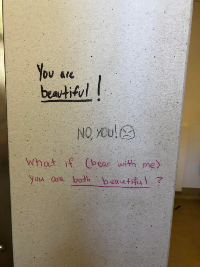 Positive and playful public property vandalism featuring compliments and a small angry face drawn on a wall.