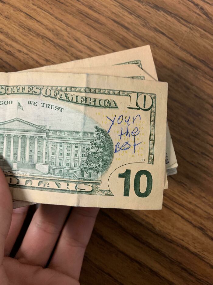 Ten dollar bill with handwritten message vandalized on public property seen in a person's hand on a wooden surface.