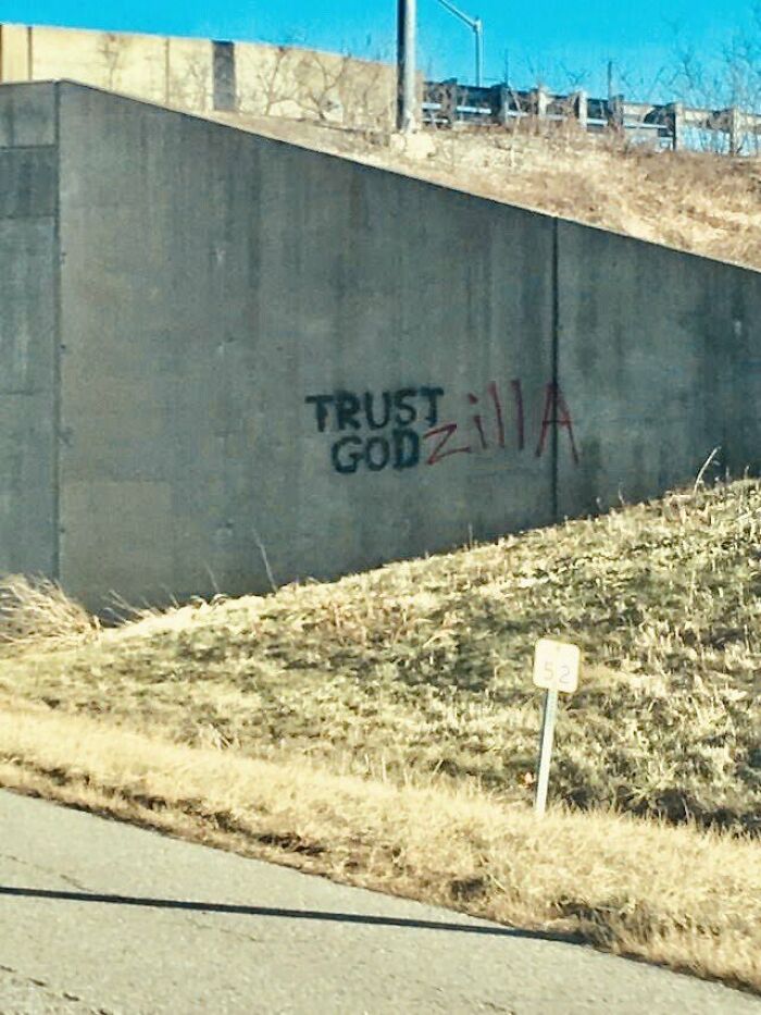 Graffiti vandalism on a concrete wall near a roadside, creatively altering the text to include Godzilla reference.