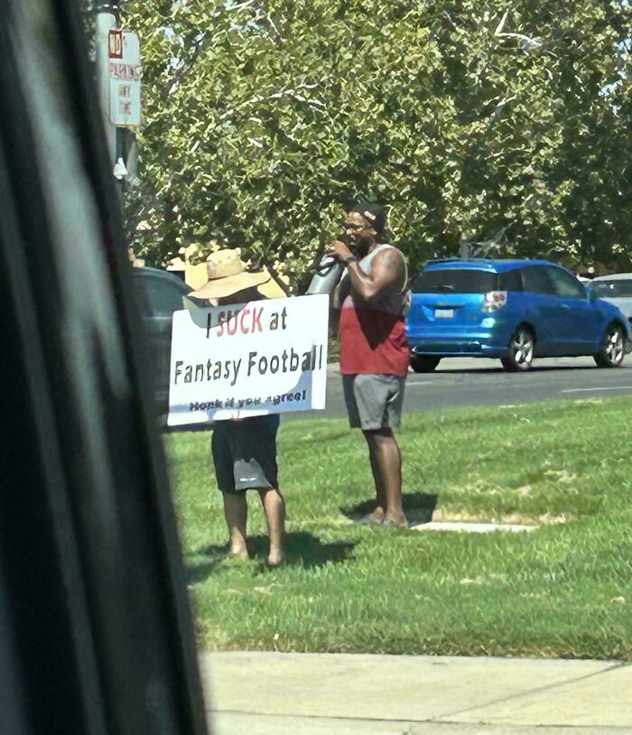 Two men standing by a roadside, one holding a humorous sign about fantasy football in a funny random notes style.