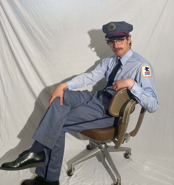 Man in a humorous uniform pose, wearing a postal worker outfit and glasses, sitting casually on an office chair.