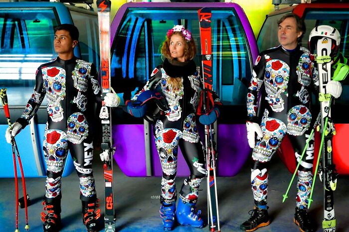 Three skiers wearing colorful skull-themed ski uniforms holding skis and poles, standing in front of gondolas.