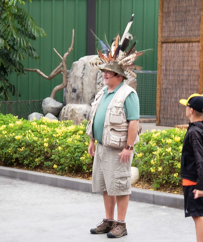 Man wearing a funny uniform with a feathered hat, beige vest, and shorts, standing outdoors near greenery and rocks.