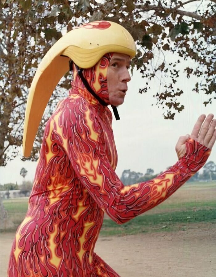 Man in a flame-patterned uniform and large novelty helmet posing outdoors, showing funny uniform moments.