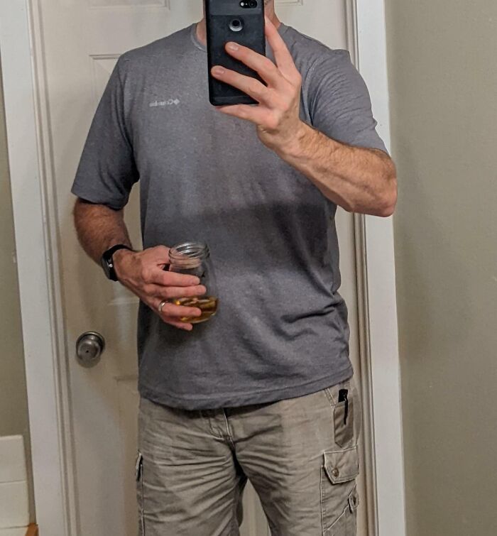 Man wearing casual uniform with grey shirt and cargo shorts taking mirror selfie holding a drink in a jar indoors