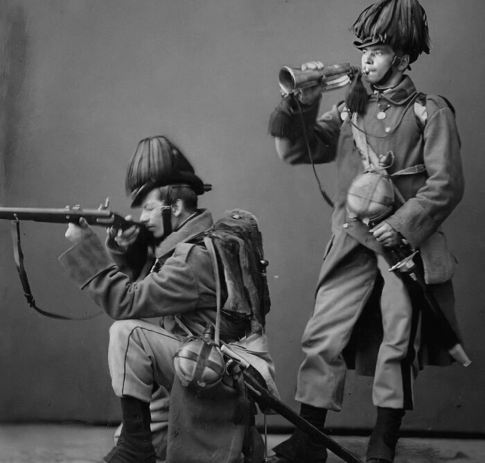 Two soldiers in historical uniforms posing with a rifle and bugle in a black-and-white vintage photo.