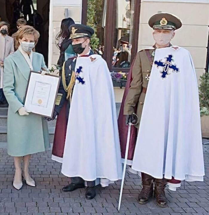 Two men in white capes and uniforms standing beside a woman holding a framed certificate outdoors during a ceremony.