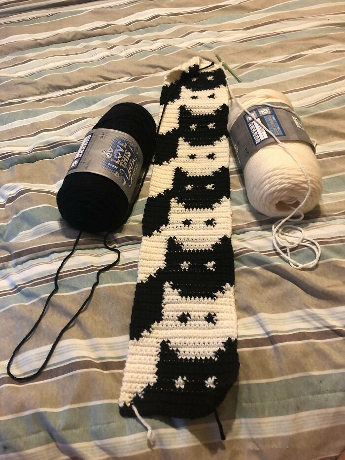 Black and white cat pattern crochet project in progress with yarn balls on a striped fabric surface.