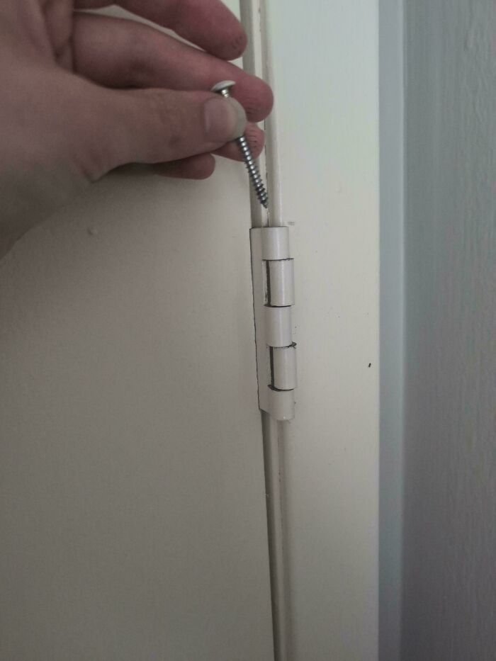 Hand holding a s***w near a door hinge with visible gaps, showing poor landlord maintenance in a rental property.