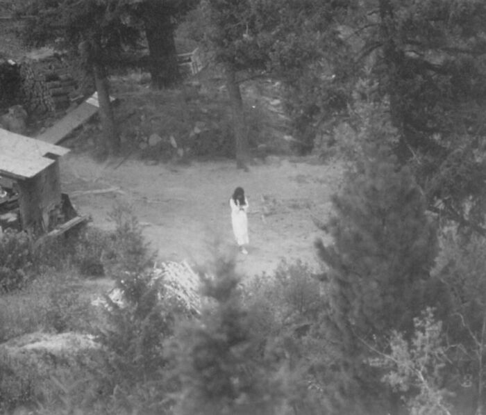Black and white eerie photo of a woman in white surrounded by trees, evoking terrifying and scary atmosphere.