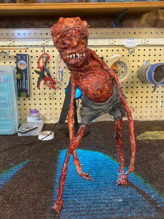 Terrifying creepy creature figure with distorted face and long limbs standing on a workshop table for scary photos collection.