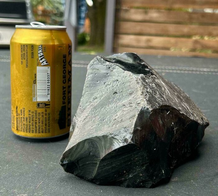 Black shiny rock with smooth surfaces placed next to a yellow soda can showing moderately interesting texture details.